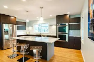 A modern kitchen featuring dark brown cabinets, stainless steel appliances, a long central island with a light marble countertop and clear acrylic bar stools, and light blue subway tile backsplash.