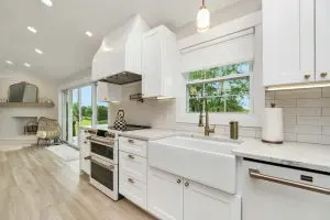 A bright, modern farmhouse kitchen with white cabinets, gold hardware, a deep farmhouse sink, marble-look countertops, and a white subway tile backsplash, overlooking a sliding glass door and the outdoors.