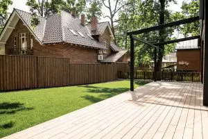 A spacious backyard with a light-colored wooden deck, a modern black metal pergola frame, a bright green lawn, and a tall wooden fence separating it from a traditional red brick house with a terracotta tile roof.