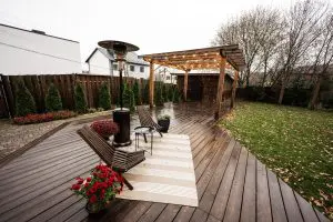 A wet outdoor patio or deck made of dark wood planks featuring a wooden pergola strung with lights, a patio heater, modern wooden chairs, and a small rug on a cloudy day.