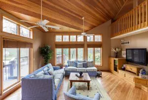 A spacious, rustic-modern living room with a high vaulted ceiling clad in natural wood, two blue floral-patterned sofas, light wood floors, and large windows looking out onto a deck and wooded area.