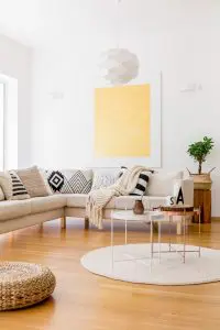 A bright, minimalist living room with a beige sectional sofa covered in black and white patterned pillows, light wooden floors, a woven round rug, and a large yellow abstract painting on the white wall.