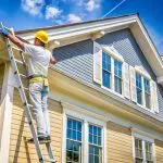 Painter on ladder working on siding to show how long does an exterior painting project take for a single-family home