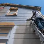 Painter on ladder applying primer to home siding, illustrating what to expect before an exterior painting project starts