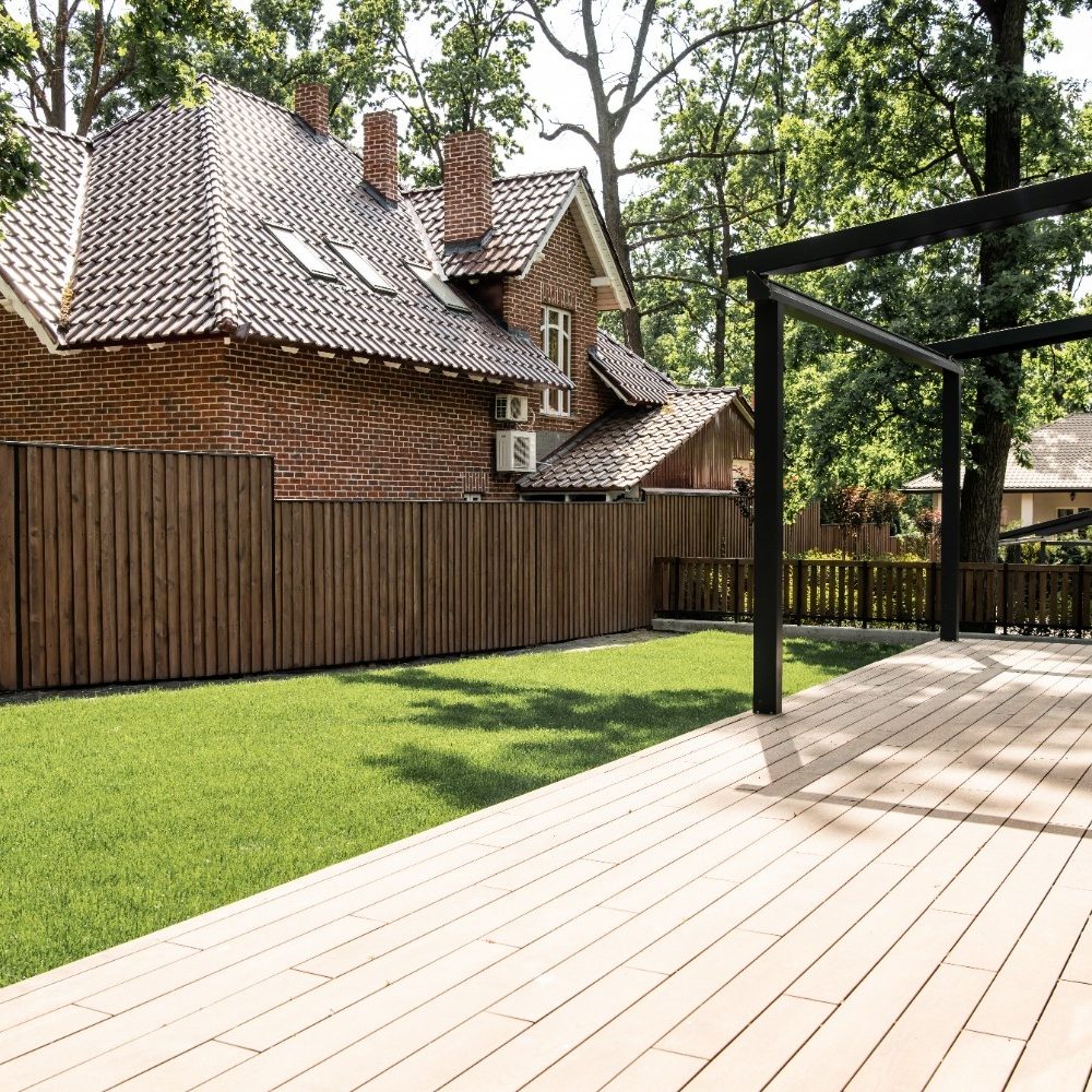 A spacious backyard with a light-colored wooden deck, a modern black metal pergola frame, a bright green lawn, and a tall wooden fence separating it from a traditional red brick house with a terracotta tile roof.
