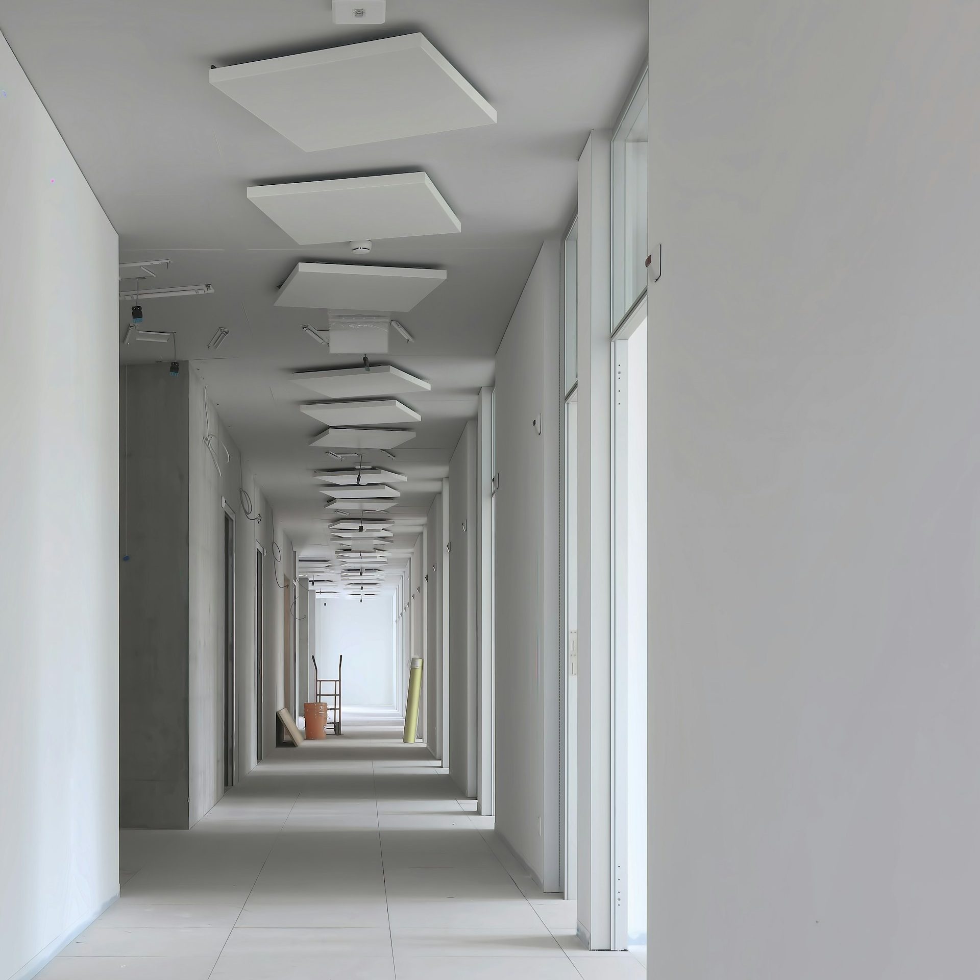 Drywall services in fort myers (3) A long, stark, and empty hallway in an unfurnished construction space, featuring plain white walls and ceiling, gray tiled floors, and acoustic ceiling panels suspended at various heights.