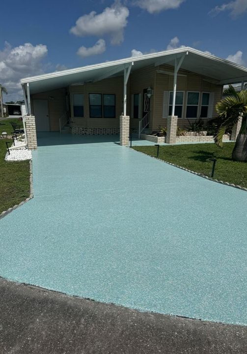 A single-story mobile home with tan siding, a white car-port roof, and a speckled light blue driveway/carport floor, bordered by a green lawn on a sunny day.