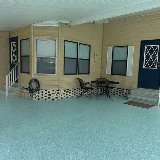 A sheltered mobile home carport/patio with tan vinyl siding, a speckled light blue floor, two dark entry doors, and a small wrought-iron table and chairs.