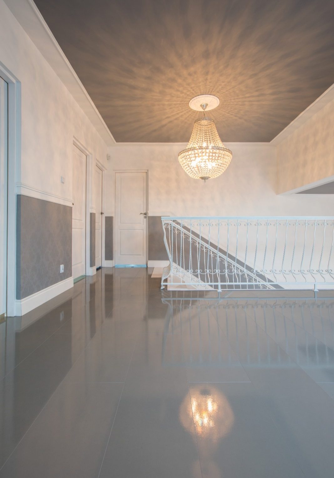A luxurious hallway and landing with a high-gloss grey floor reflecting a crystal chandelier hanging from a dark grey ceiling with dramatic light patterns, contrasting with white walls and a white metal balustrade.