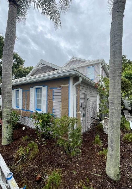 A gray-sided house under renovation, with windows taped off and protected by paper and blue painter's tape, surrounded by landscaping, mulch, and two large palm trees in the foreground.