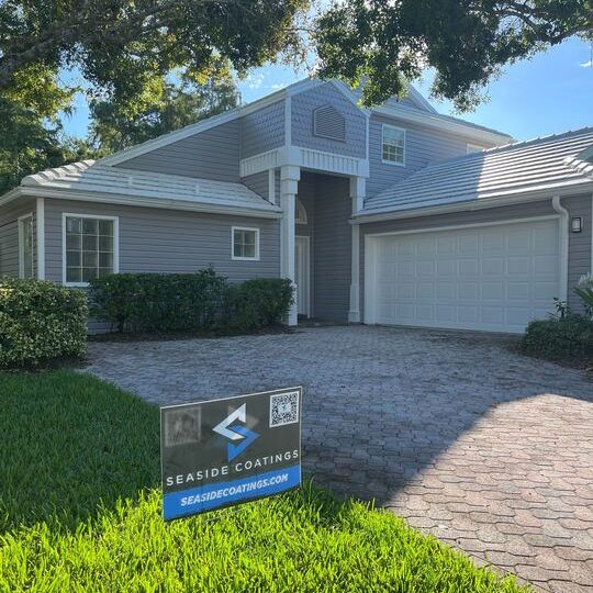 A two-story house with light gray siding, a white garage door, a tile roof, and a paver driveway, with a Seaside Coatings sign on the bright green lawn.