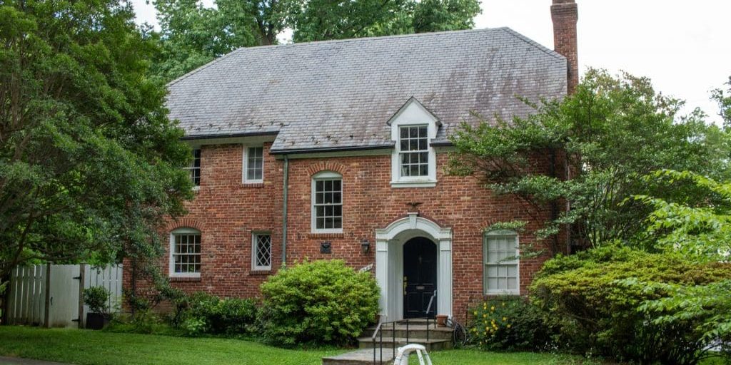 A traditional **two-story red brick house** with a gray slate roof, white-trimmed windows, a chimney, and a small white toy car parked on the front walk.
