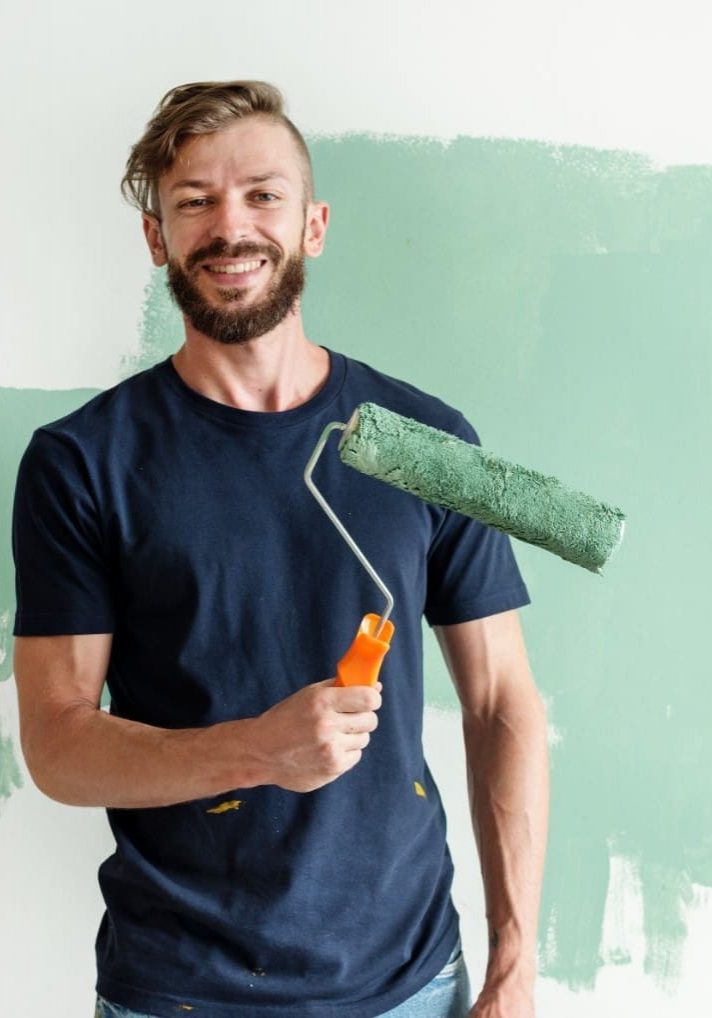 A man with a beard and a dark shirt holding a paint roller with green paint in front of a partially painted wall.