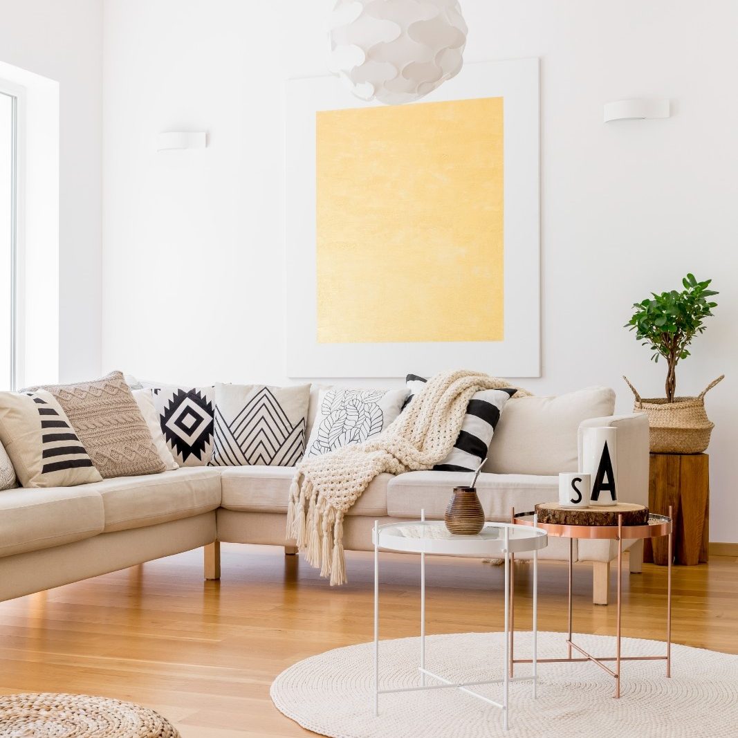 A bright, minimalist living room with a beige sectional sofa covered in black and white patterned pillows, light wooden floors, a woven round rug, and a large yellow abstract painting on the white wall.