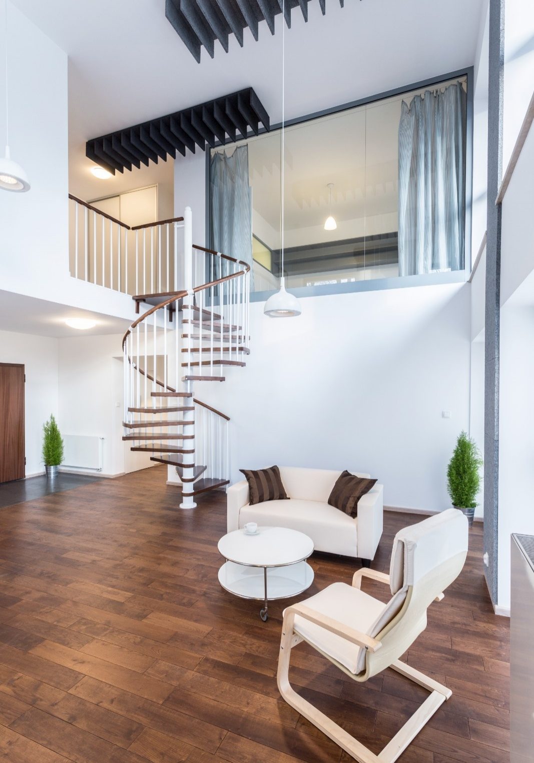 A bright, modern loft living room with dark wood flooring, a white spiral staircase leading to an upper level with a large window, a small white sofa, and a modern lounge chair.