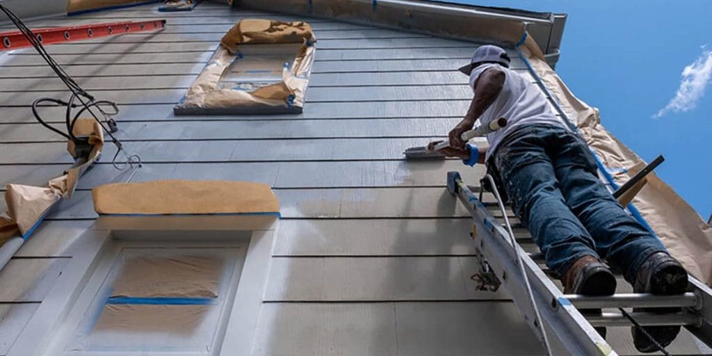 Painter on ladder applying primer to home siding, illustrating what to expect before an exterior painting project starts